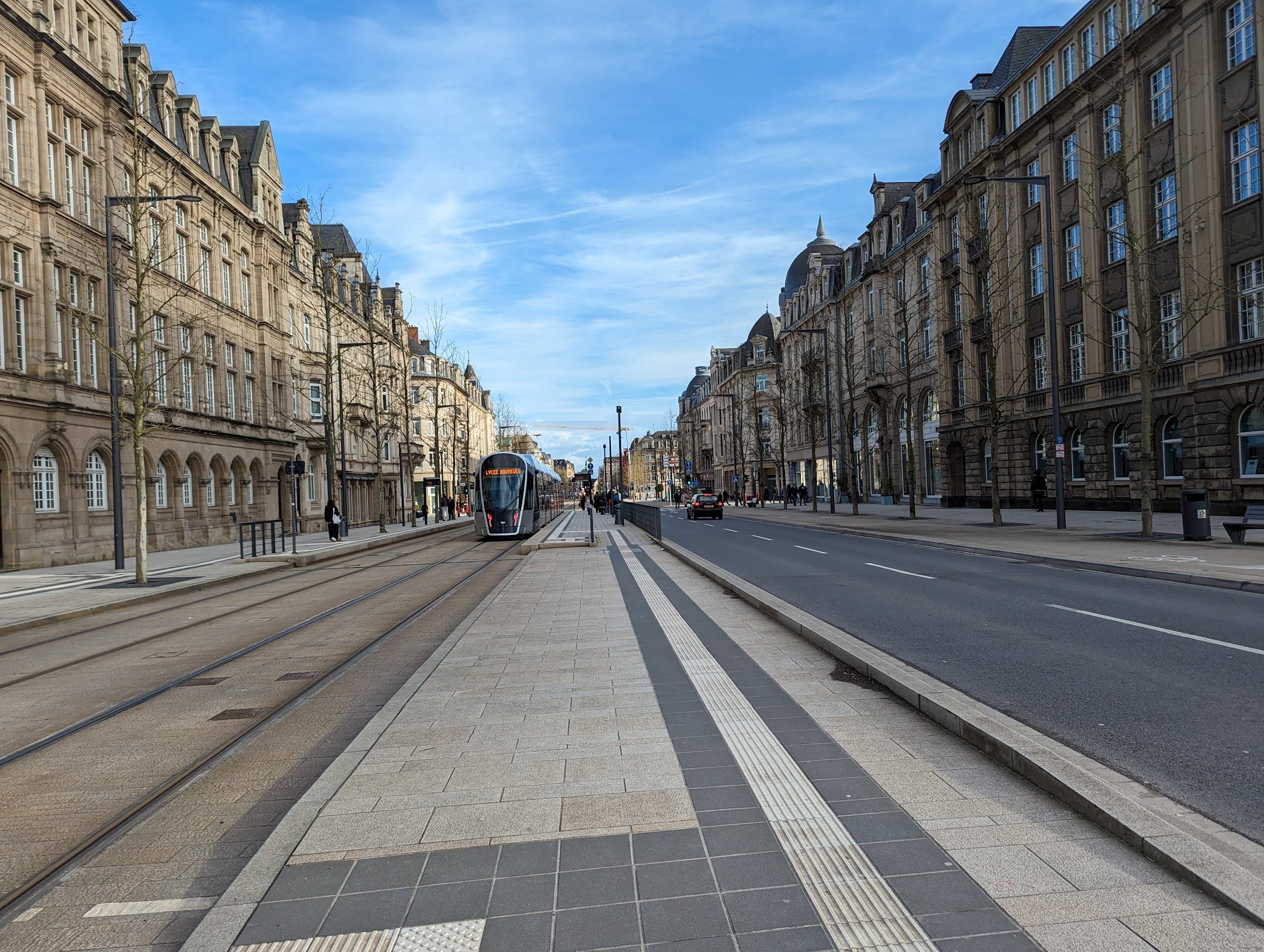 A photo I took standing in the middle of one of the main streets in Luxembourg. On either side are shops of about 4 floors, which are a maybe 2/5 the height of the width of the street. There are quite wide pavements, and on the left two trams lanes and a tram in shot, and more pavement and tramstops in the centre. To the right are two reasonably narrow road lanes and to its right a cycle path. The buildings and street (although not the road) are yellow-grey and well-kept, with trees, benches, and bins.